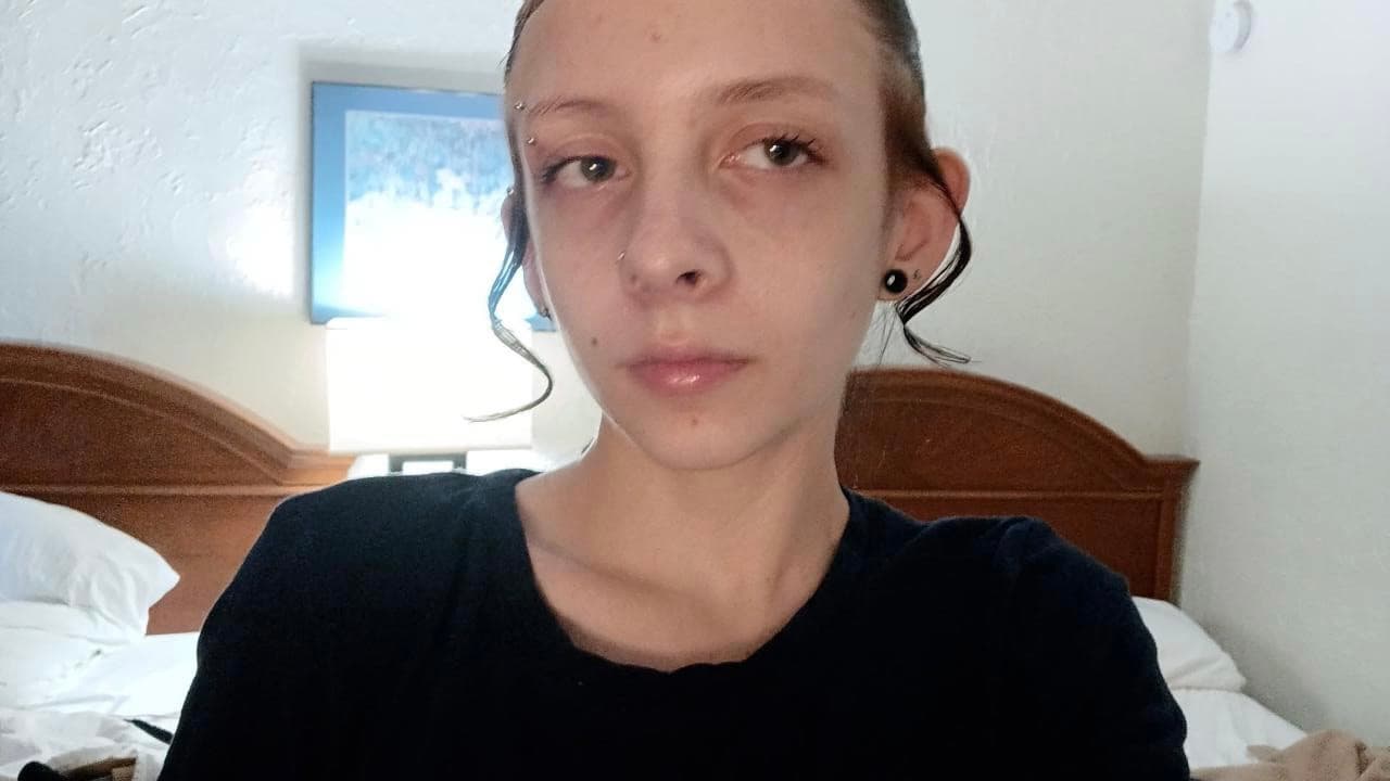 Person with facial piercings and dark hair curls, wearing a black shirt in a bedroom.