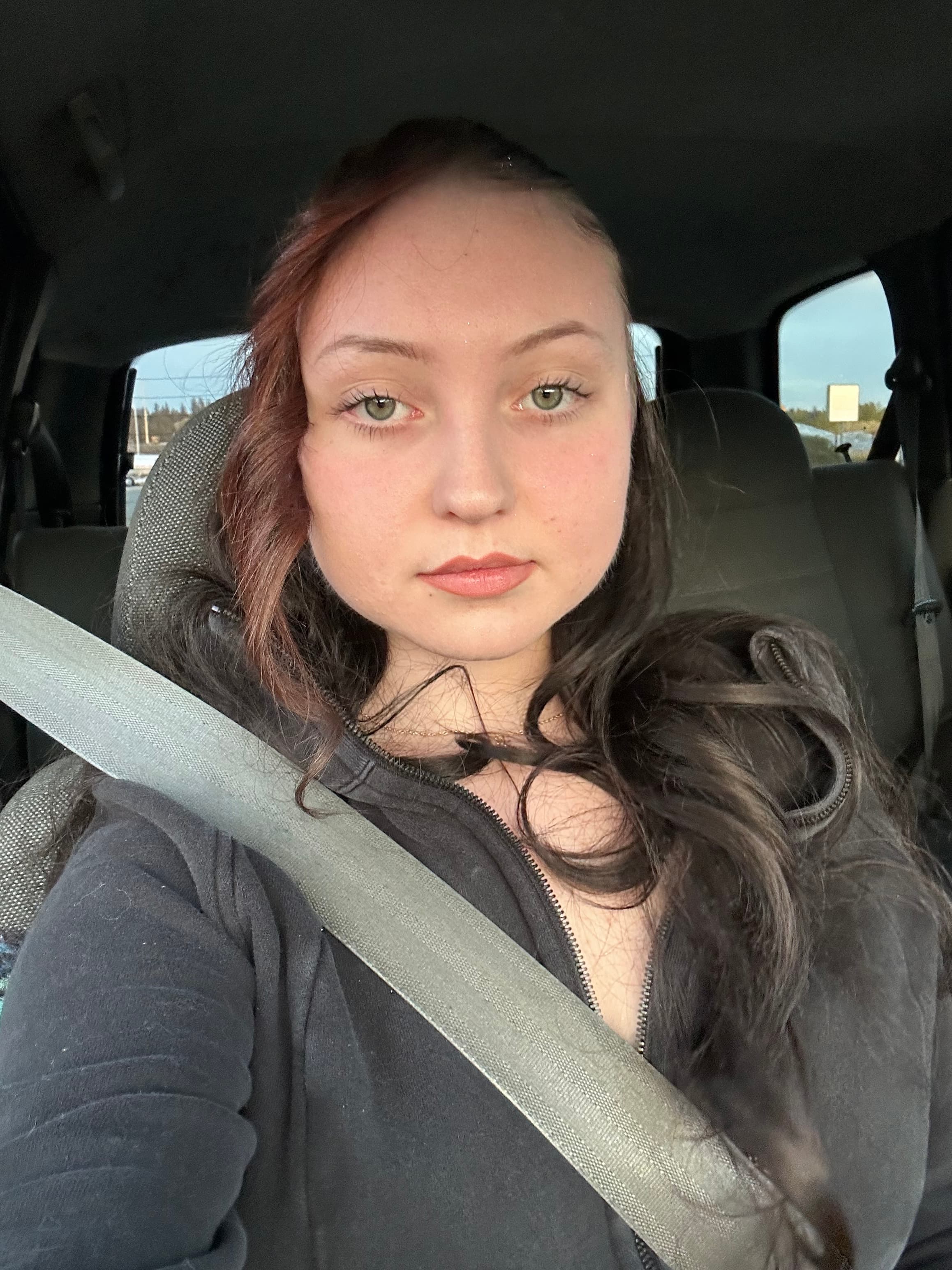 Young woman with green eyes and long wavy hair wearing a seatbelt in a car.