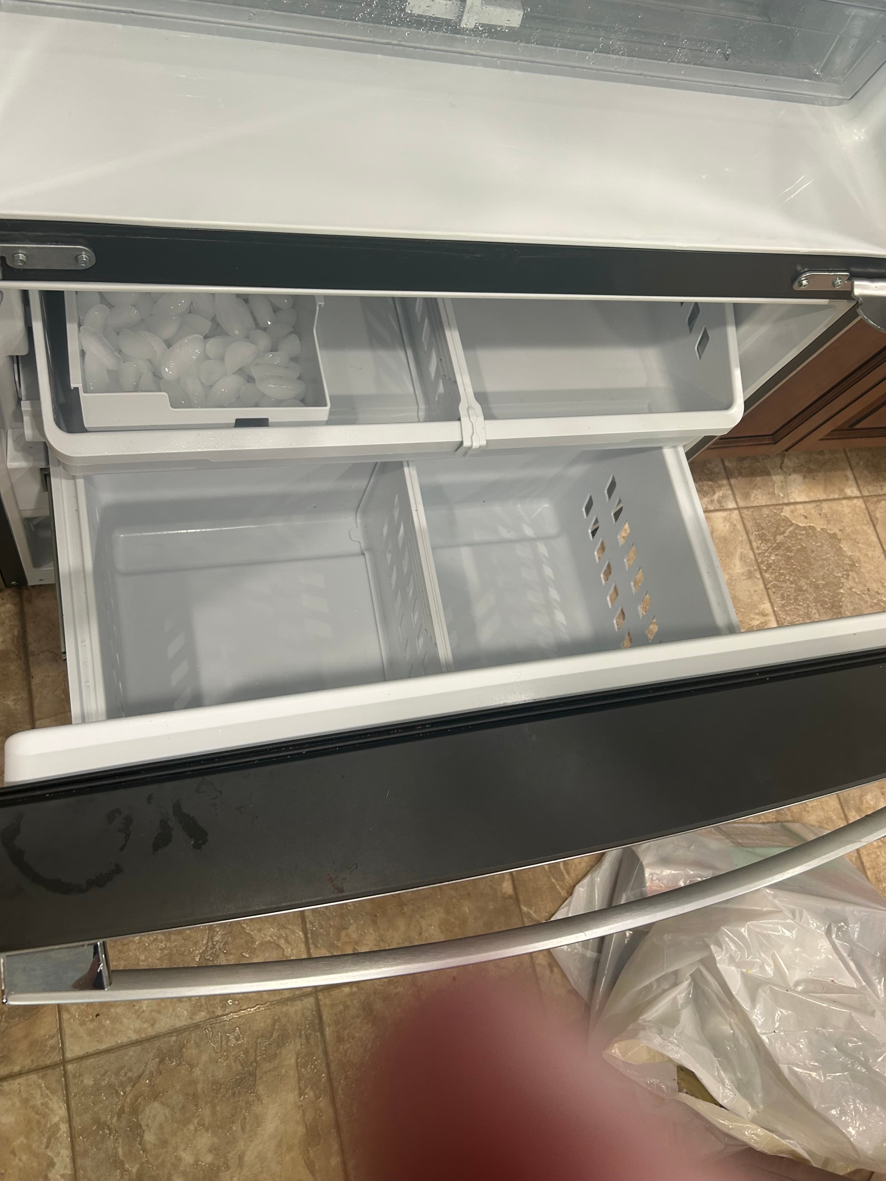 Open refrigerator freezer drawer with ice cubes in a white bin and several empty compartments.